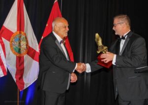 Two men shaking hands during an award ceremony with flags in the background.
